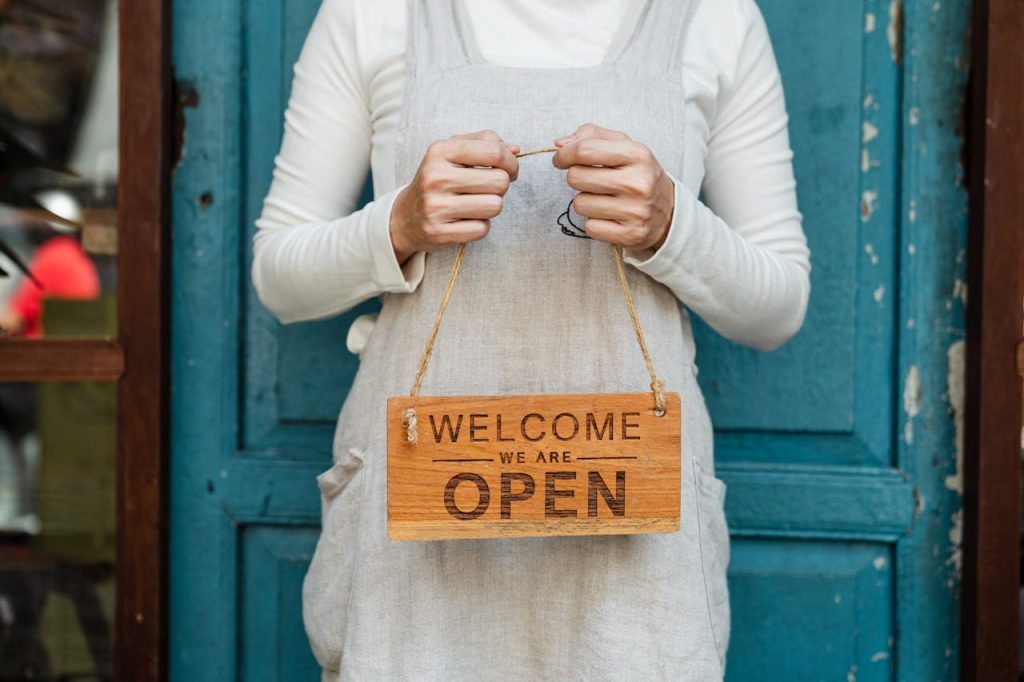 A person holds a Welcome, we are open sign in front of a rustic blue door.